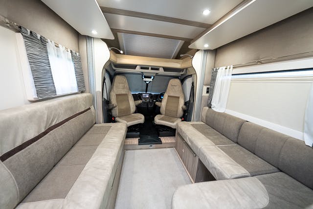 Interior view of a 2019 Benimar Tessoro T486 motorhome featuring two beige captain chairs at the front, beige and grey upholstered sofas on both sides, a neutral-toned floor, and small windows with white curtains. The space is clean and well-lit with built-in ceiling lights.