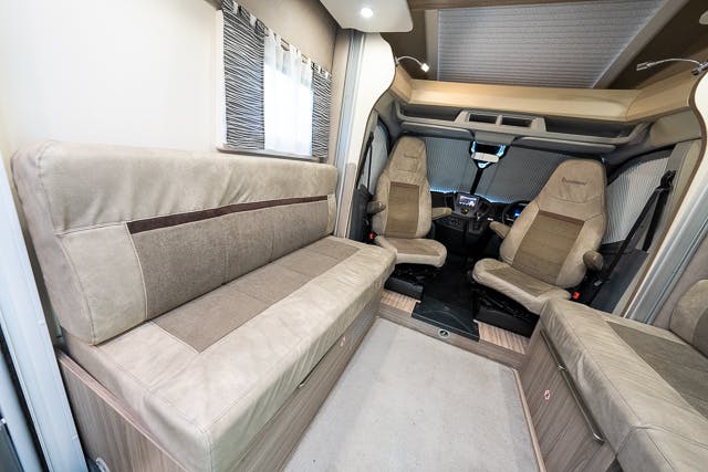 Interior view of a 2019 Benimar Tessoro T486 camper van showing the driver's cabin with two seats and a dashboard, two cushioned benches opposite each other, and a window with patterned curtains on the left wall. The interior features beige and brown tones with wooden fixtures.