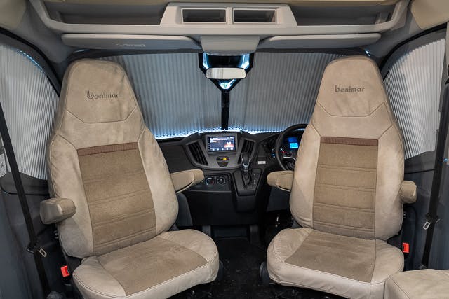 The image shows the interior cabin of a 2019 Benimar Tessoro T486 vehicle, featuring two beige upholstered seats with armrests. A dashboard with a central display screen and steering wheel is visible, along with a corrugated sunshield on the front windshield.