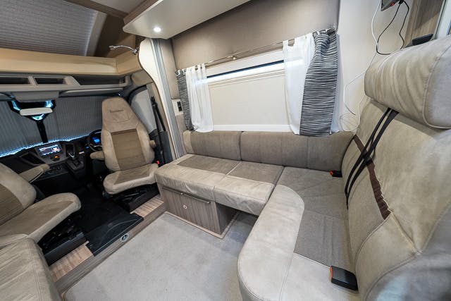 Interior view of the 2019 Benimar Tessoro T486 RV showing a seating area with beige upholstery. The layout includes swivel driver and passenger seats, a U-shaped sofa, a window with white curtains, and a pull-down blind. The space is well-lit and neatly arranged.