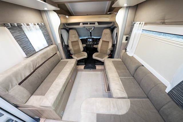 The 2019 Benimar Tessoro T486 motorhome showcases a chic interior with beige and brown upholstery, featuring two long sofas facing each other, a compact table, and a view toward the front driver's area with two swivel seats. The space is well-lit with white curtains on the windows.