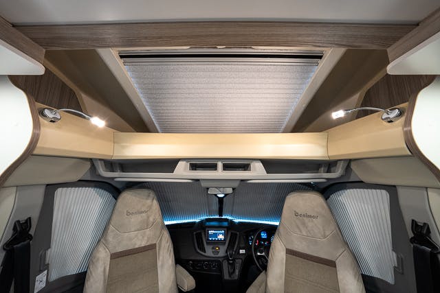 The interior of the 2019 Benimar Tessoro T486 motorhome is shown, focusing on the front area with two upholstered seats, a dashboard with a steering wheel, digital displays, and overhead storage compartments. The space is well-lit with built-in lights.