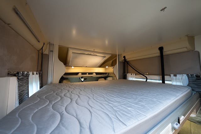 Interior view of a small bedroom area in the 2019 Benimar Tessoro T486 RV. The image features a raised, neatly-made bed with a quilted mattress, surrounded by curtains on the sides and windows at the back. The lighting is warm, and the space appears cozy.