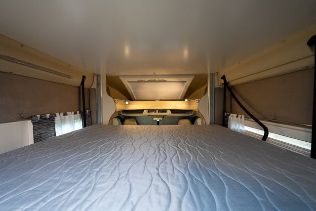Wide-angle view of the interior of a 2019 Benimar Tessoro T486 campervan showing a bed with a gray quilted mattress cover. The bed is positioned over the driver's cabin area. The space is surrounded by beige walls, and small windows with curtains are partially visible on the sides.