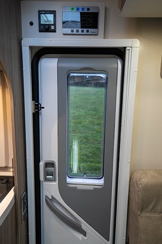 Interior view of the 2019 Benimar Tessoro T486 RV door with a window. A control panel is mounted above the door, showing various indicators and buttons. Part of a kitchen counter and sink is visible on the left, and a portion of a beige seat is visible on the right.