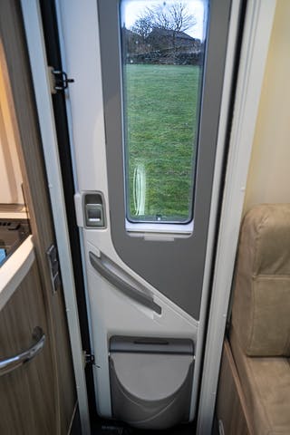 Interior view of the 2019 Benimar Tessoro T486 motorhome door with a vertical window. The door has a latch and handle on the left side, with additional storage compartments integrated into the lower section. The scene outside reveals a grassy area and an overcast sky.