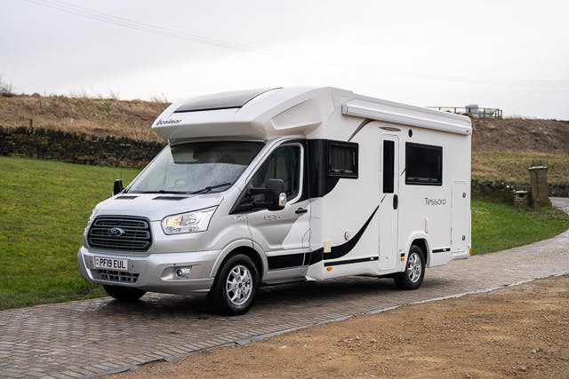 A white 2019 Benimar Tessoro T486 motorhome is parked on a brick driveway, surrounded by grassy and stone areas. The motorhome features a "Trigano" logo and a UK license plate "PF19 EUL." The rural setting includes a fence and trees in the background.