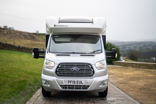 A white 2019 Benimar Tessoro T486 motorhome with "Benimar" branding on the front, parked on a paved and grassy area. The license plate reads "PF19 EUL." The background includes a blurred view of a rural landscape with rolling hills and overcast skies.