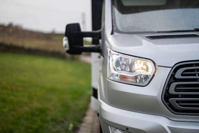 A close-up shot of the right front side of a 2019 Benimar Tessoro T486 silver van. The image highlights the front headlight, which is turned on, and part of the front grille. The background shows a grassy area and a blurred out-of-focus landscape.