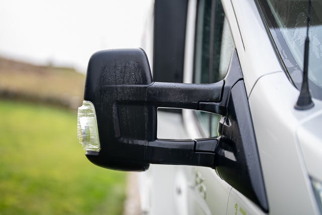 Close-up image of a black, rectangular side mirror attached to a white 2019 Benimar Tessoro T486. The mirror is wet, likely from rain, and has an integrated turn signal indicator. The background is slightly blurred, showing a grassy area.