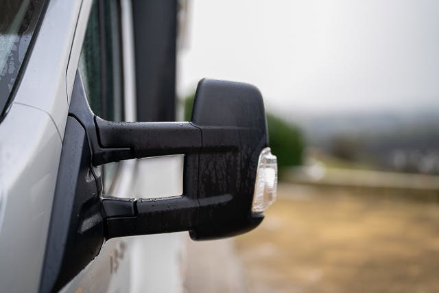 Close-up of a black side view mirror on a 2019 Benimar Tessoro T486. The mirror is wet, suggesting recent rain. The blurred background shows an out-of-focus landscape with grassy and distant elements. The focus is on the detail of the mirror and the vehicle's side.