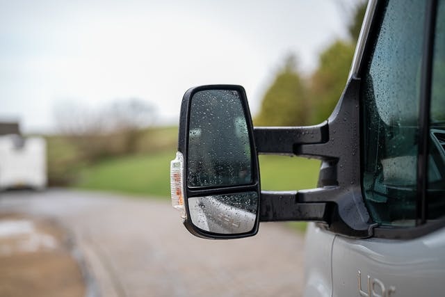 A close-up shot of a side mirror on a 2019 Benimar Tessoro T486 displaying raindrops on its surface. The background features a blurred outdoor setting with greenery and a cobblestone area. The edge of the vehicle is partially visible on the right side of the image.
