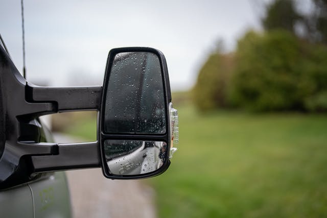 A close-up shot of a 2019 Benimar Tessoro T486's side view mirror, wet from rain. The mirror reflects part of the vehicle and a blurry view of the grassy landscape and trees in the background. The weather appears overcast.