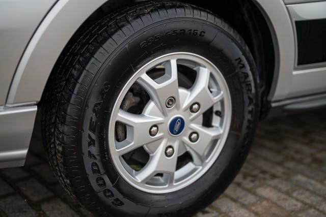 Close-up view of a silver car's tire and wheel on a paved surface. The tire is a Goodyear brand, with the model name "Marathon" visible on the sidewall. The wheel, reminiscent of a 2019 Benimar Tessoro T486, features a multi-spoke design with a central blue and silver logo.