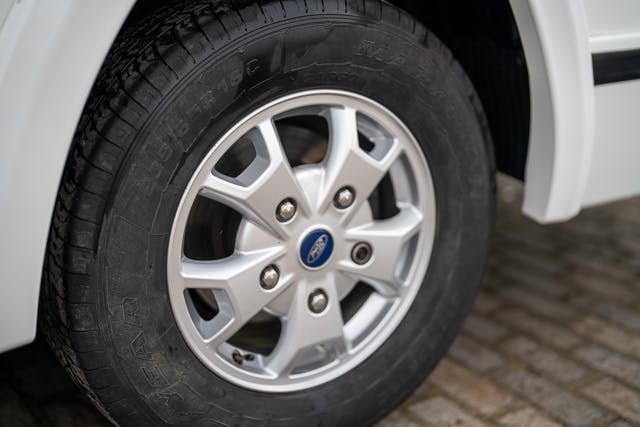 Close-up view of a car's front wheel featuring a five-spoke alloy rim with a Ford logo in the center, reminiscent of the design aesthetics seen in models like the 2019 Benimar Tessoro T486. The tire has visible treads and markings, set against a ground paved with interlocking bricks.