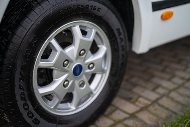 Close-up image of a 2019 Benimar Tessoro T486's front wheel featuring a silver alloy rim and Goodyear tire with the specification 235/65 R16C. The wheel is mounted on a white vehicle, and the car is parked on a paved surface.