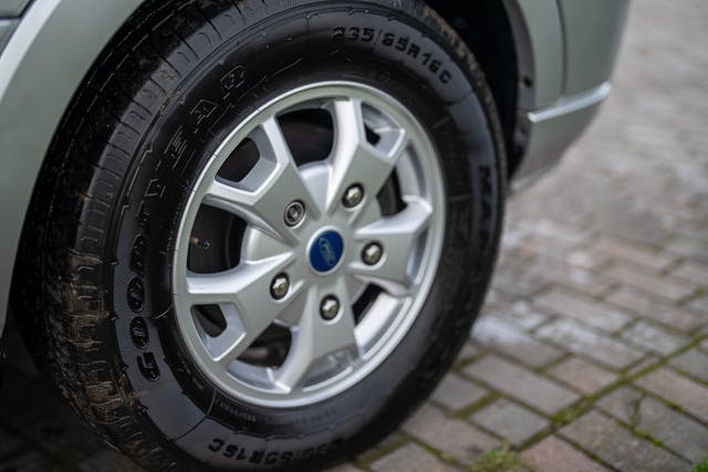 Close-up shot of a car's front wheel with a Goodyear tire and a silver alloy rim. The tire specifications are visible on the tire's sidewall. The 2019 Benimar Tessoro T486 is parked on a cobblestone surface, with some grass visible between the stones.