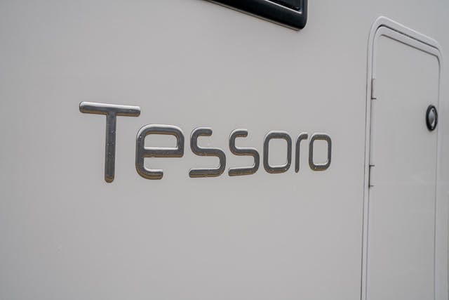 The image shows the word "Tessoro" displayed in metallic letters on the side of a pale-colored 2019 Benimar Tessoro T486. The letters are in a modern, rounded font, and the background features a closed door with a small black handle and hinge.