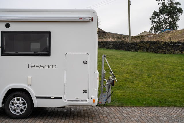The image shows the side view of a white 2019 Benimar Tessoro T486 motorhome labeled "Tessoro." The motorhome is parked on a cobblestone surface, adjacent to a grassy area with a stone wall and trees in the background. A metal bike rack is attached to the rear of the motorhome.
