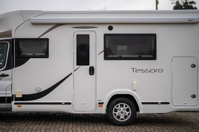 Side view of a white 2019 Benimar Tessoro T486 RV with the brand name "Tessoro" visible. The RV is parked on a cobblestone surface and features a side door, a large window, and storage compartments. An extendable awning is mounted above the door and window.
