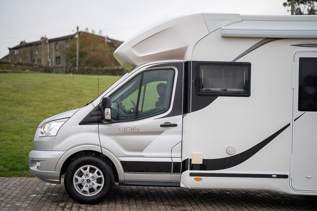 A side view of a 2019 Benimar Tessoro T486 motorhome parked on a paved surface. The vehicle features a driver's side window, door, and a window on the side of the living quarters area. A grassy field and a building are visible in the background.