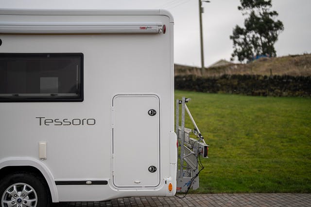 Side view of a 2019 Benimar Tessoro T486 white campervan with the word "Tessoro" on its side. The van has a small window and storage compartment visible. A bike rack is attached to the rear. The background features a grassy area, a stone wall, and an overcast sky.