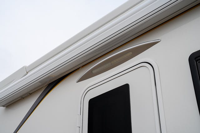 A close-up of a section of a 2019 Benimar Tessoro T486 RV. The image shows the top part of a white door, a black window, and an extended awning above. There's also a triangular decorative element above the door.