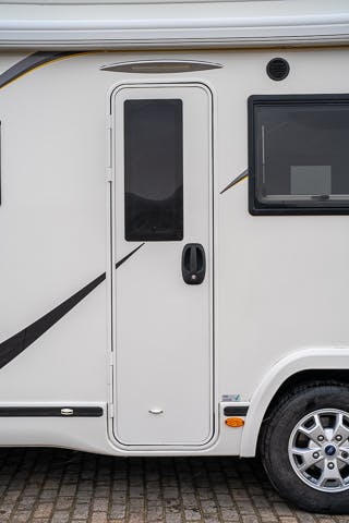 Side view of a white 2019 Benimar Tessoro T486 motorhome showing a door with a window and a black handle. To the right of the door is a rectangular window. The motorhome is parked on a cobblestone surface, and part of the rear wheel and tire is visible at the bottom.