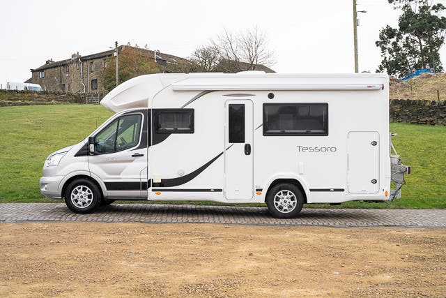 A 2019 Benimar Tessoro T486 motorhome labeled "Tessoro" is parked on a paved surface in front of a grassy area. The vehicle has multiple windows and a door on the side. In the background, there are buildings and some trees.