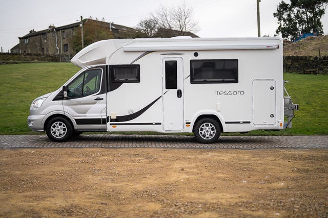 A 2019 Benimar Tessoro T486 camper van is parked on a paved surface with a grassy area and buildings in the background. The white camper van features dark windows and a sleek design, and is parked in a seemingly rural setting.