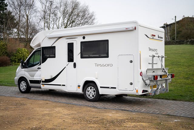 A white 2019 Benimar Tessoro T486 motorhome is parked on a paved area next to a grass field. The vehicle has several windows, a side door, and a bike rack on the rear. Trees and utility poles are visible in the background.