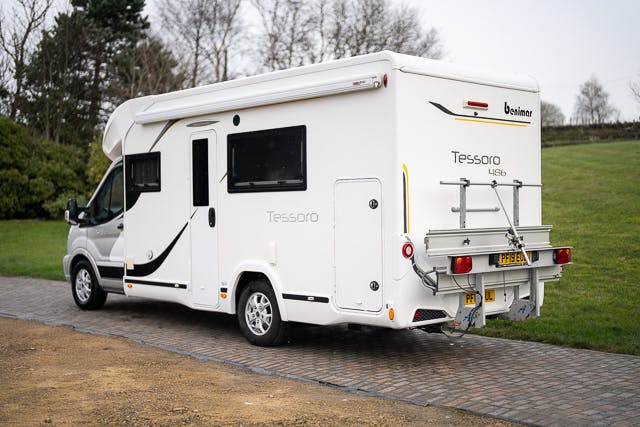 A 2019 Benimar Tessoro T486 motorhome in white is parked on a paved area next to a grassy field. The vehicle features a rear bike rack and several side compartments. Surrounding the motorhome are shrubs and leafless trees in the background.