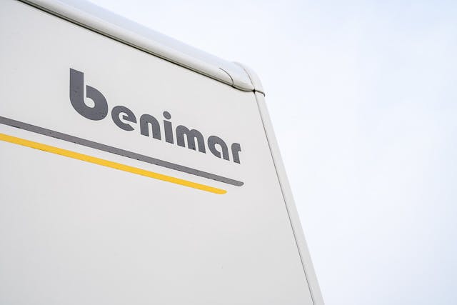 Partial view of a 2019 Benimar Tessoro T486 camper van with the logo "benimar" in gray letters on the side. A yellow and gray stripe runs horizontally below the logo. The sky in the background is overcast.