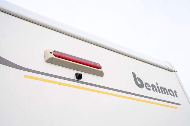 The image shows a close-up of the back of a white 2019 Benimar Tessoro T486 RV with the brand name "benimar" written on it. There's a red brake light centered above a small black camera, and a yellow stripe extends horizontally across the vehicle.
