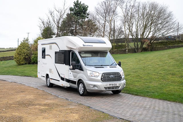 A white 2019 Benimar Tessoro T486 motorhome is parked on a paved pathway with grass on either side. The vehicle has a front grille with the Ford logo and a license plate reading "PF18 EUL". There are trees in the background, and the sky is overcast.