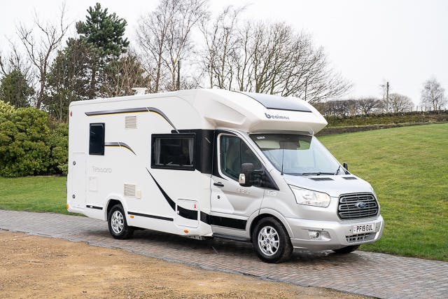 A 2019 Benimar Tessoro T486 motorhome with a Ford Transit base is parked on a stone driveway. The vehicle features black and gold detailing on its sides, large windows, and various compartments. Surrounding the driveway are trimmed hedges and bare trees, indicating a winter setting.