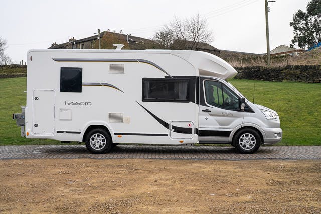 A 2019 Benimar Tessoro T486 motorhome is parked on a paved driveway. The vehicle has a streamlined design with graphics on the side and multiple windows. A grassy area and stone wall are visible in the background.