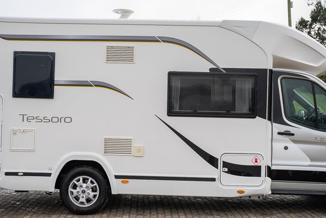 Side view of a white 2019 Benimar Tessoro T486 motorhome with "Tessoro" written on the side. It features a window, a vent, and a door panel. The motorhome has black and yellow graphics and is parked on a paved surface. Part of the cab's front section is visible on the right side of the image.