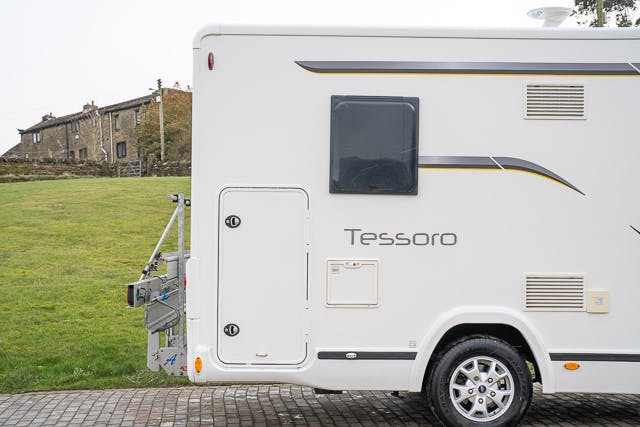 A white 2019 Benimar Tessoro T486 camper van is parked on a paved area next to a grassy field. Part of an old stone building is visible in the background to the left. The side of the camper shows a window, a door, and some storage compartments.