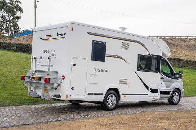 A 2019 Benimar Tessoro T486 motorhome is parked on a paved surface next to a grassy area. The vehicle has a bike rack mounted on the back and features a few small windows and vents on its side. The motorhome model name, Tessoro 486, is visible on the rear.
