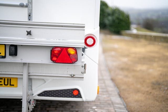 Close-up of the rear of a 2019 Benimar Tessoro T486 RV parked on a paved driveway, showing its taillights and license plate. The surrounding area is blurred, with dry grass visible on the side. The image is taken during a cloudy day.