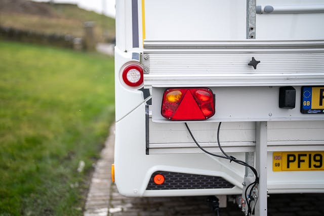 A close-up of the rear section of a white 2019 Benimar Tessoro T486 trailer. The image shows tail lights, a license plate with the characters "PFI9," and several cables connected to the back. The trailer is parked on the right side of a cobblestone path with grass on the left.