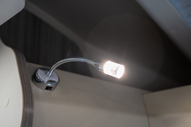 A close-up view of a lit gooseneck LED reading lamp with a flexible silver neck attached to the wall inside the 2019 Benimar Tessoro T486. The background appears to be part of the RV interior with muted colors, creating a cozy ambiance for nighttime reading.
