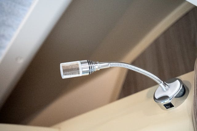 Close-up of a flexible, silver, gooseneck reading light mounted on a beige surface within the 2019 Benimar Tessoro T486. The light extends outward and features a small cylindrical LED lamp head. The surrounding area includes a partial view of a smooth, wooden texture.
