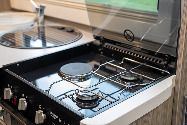 This image shows a small kitchen area inside a 2019 Benimar Tessoro T486 camper van. It features a two-burner gas stove with a glass cover, a round sink with a faucet, and a window above the stove. The stove has knobs for controlling the burners.