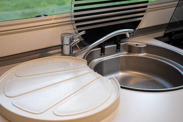 A stainless steel kitchen sink with a modern chrome faucet is partially covered by a white plastic cutting board with a cut-out section. The setup appears to be in the compact kitchen of a 2019 Benimar Tessoro T486, likely within a mobile home or RV.