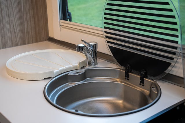 A small stainless steel sink with a tap is shown in an RV kitchen of the 2019 Benimar Tessoro T486. The sink has two lids, one closed and one open. The white countertop contrasts with a window in the background, offering a view of green grass.