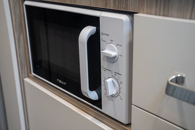 A 2019 Benimar Tessoro T486 microwave oven with a white exterior and black front is installed in a wooden and white kitchen cabinet. It has two control knobs; one for power levels and the other for time settings. The door features a white handle.