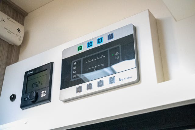 A close-up view of two electronic control panels mounted on a wall in the 2019 Benimar Tessoro T486. The left panel displays a time reading of 05:21 and has a few buttons with symbols. The right panel features a larger screen with several icons and buttons labeled "Benimar.