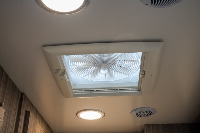 A ceiling in the 2019 Benimar Tessoro T486 features a white rectangular ventilation fan, two round ceiling lights, and a small round vent. The walls visible in the image are wooden with a light brown color.